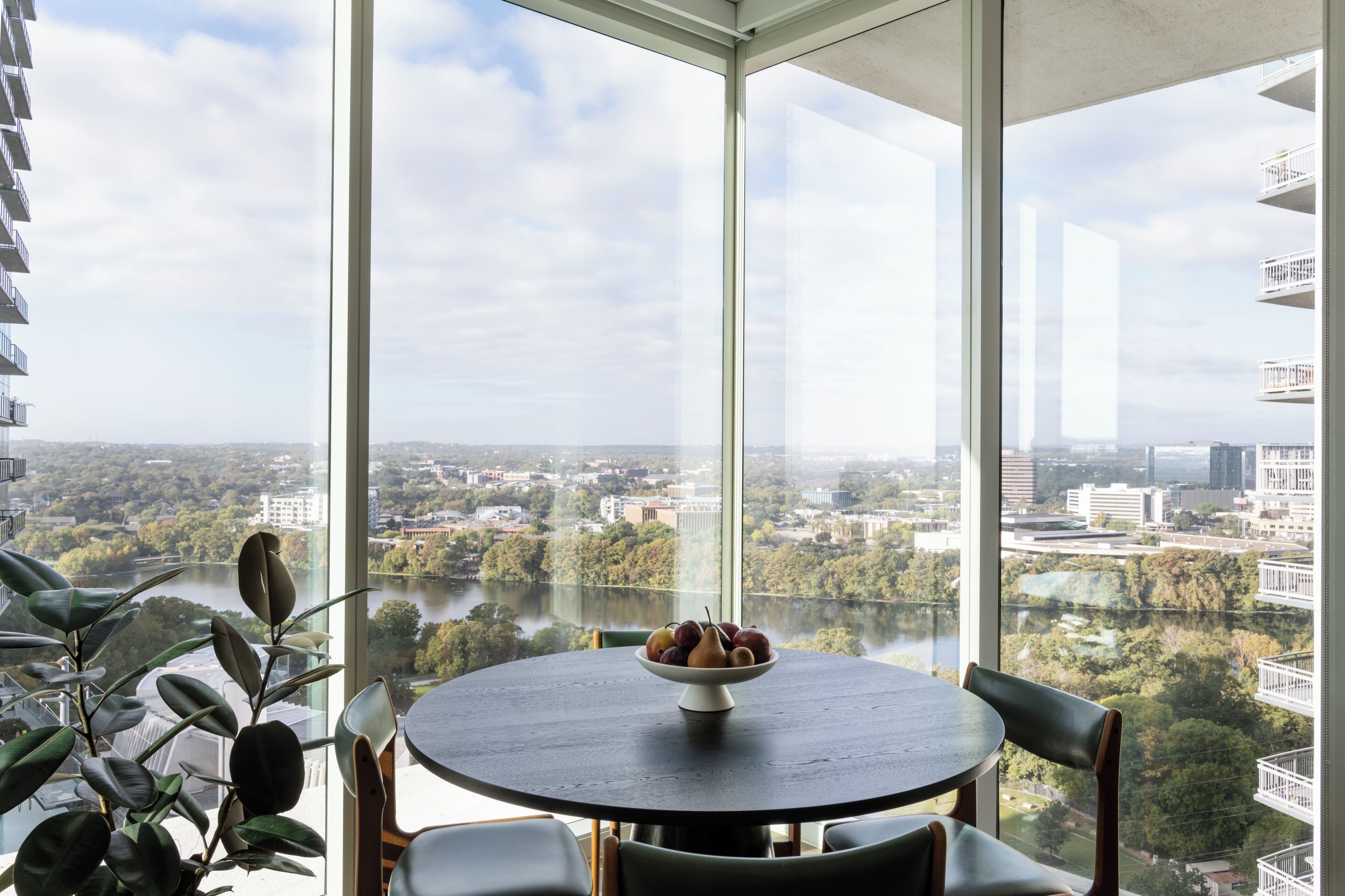 A round table with a fruit bowl, a chair, and a view of a river and cityscape through large windows.