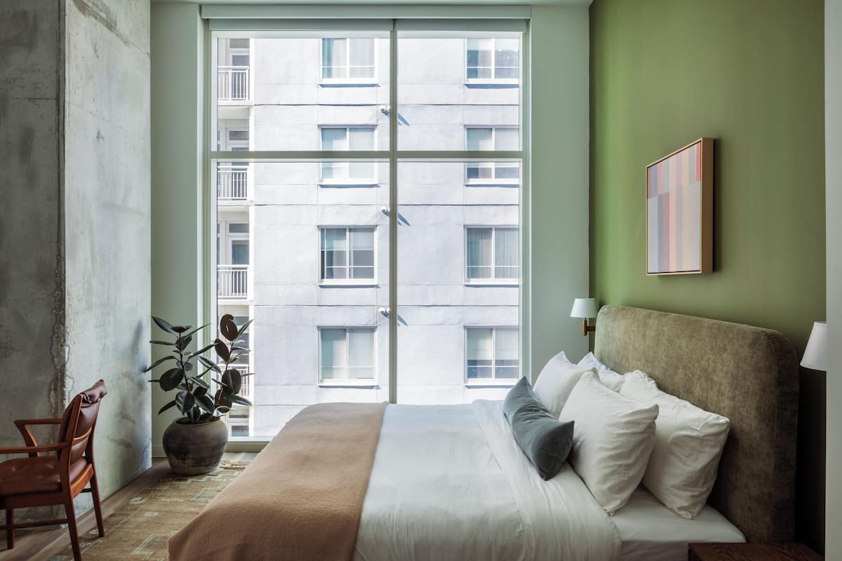 Un dormitorio con un ventanal grande, una cama con sábanas blancas, una manta marrón, una planta en maceta y un cuadro en la pared.