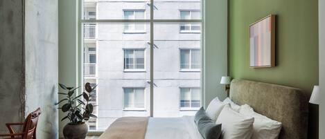 A bedroom with a large window, a bed with white linens, a brown throw blanket, a potted plant, and a framed picture on the wall.