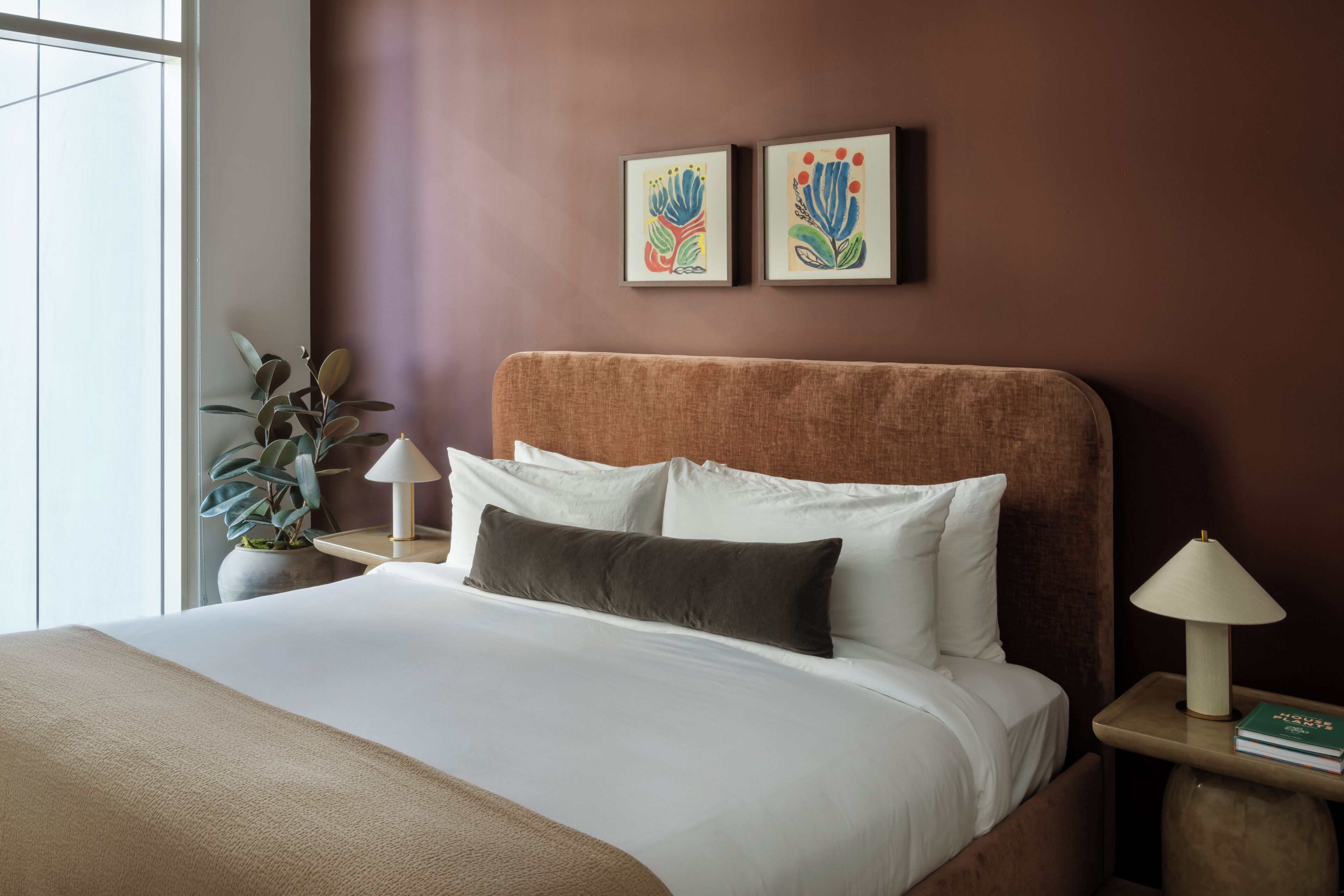 Una habitación de hotel con una cama, una mesita de noche, una lámpara, una planta en maceta y dos cuadros en la pared.