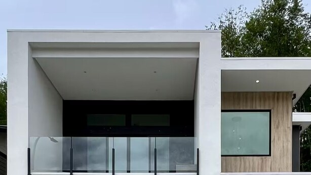 Une maison moderne à deux étages, dotée d’un garage, d’un balcon avec des garde-corps en verre et d’une grande fenêtre à l’étage supérieur.