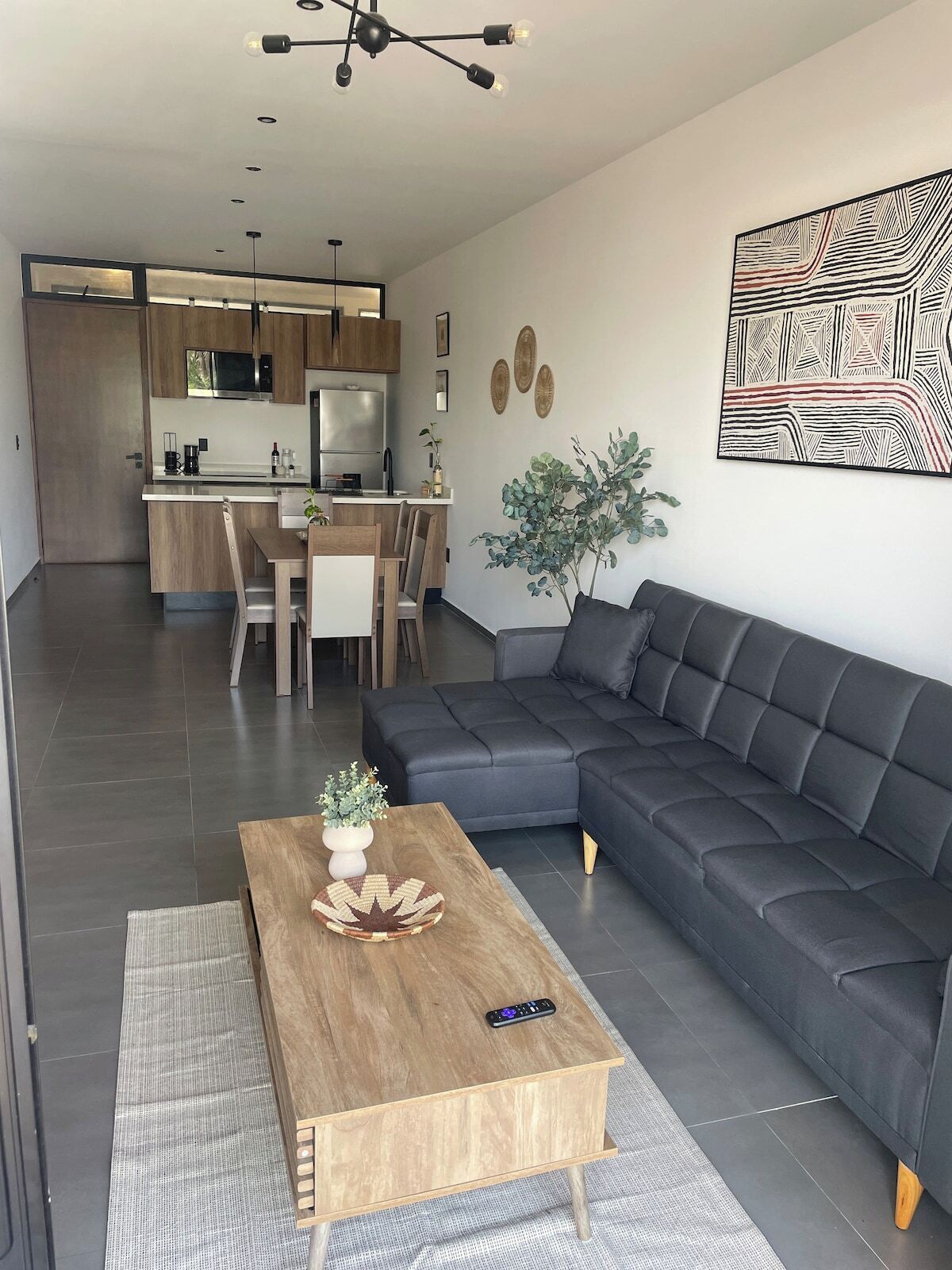 A modern living room with a dark grey sofa, a wooden coffee table with a vase and remote, and a kitchen in the background.