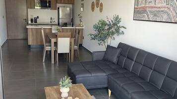 A modern living room with a dark grey sofa, a wooden coffee table with a vase and remote, and a kitchen in the background.