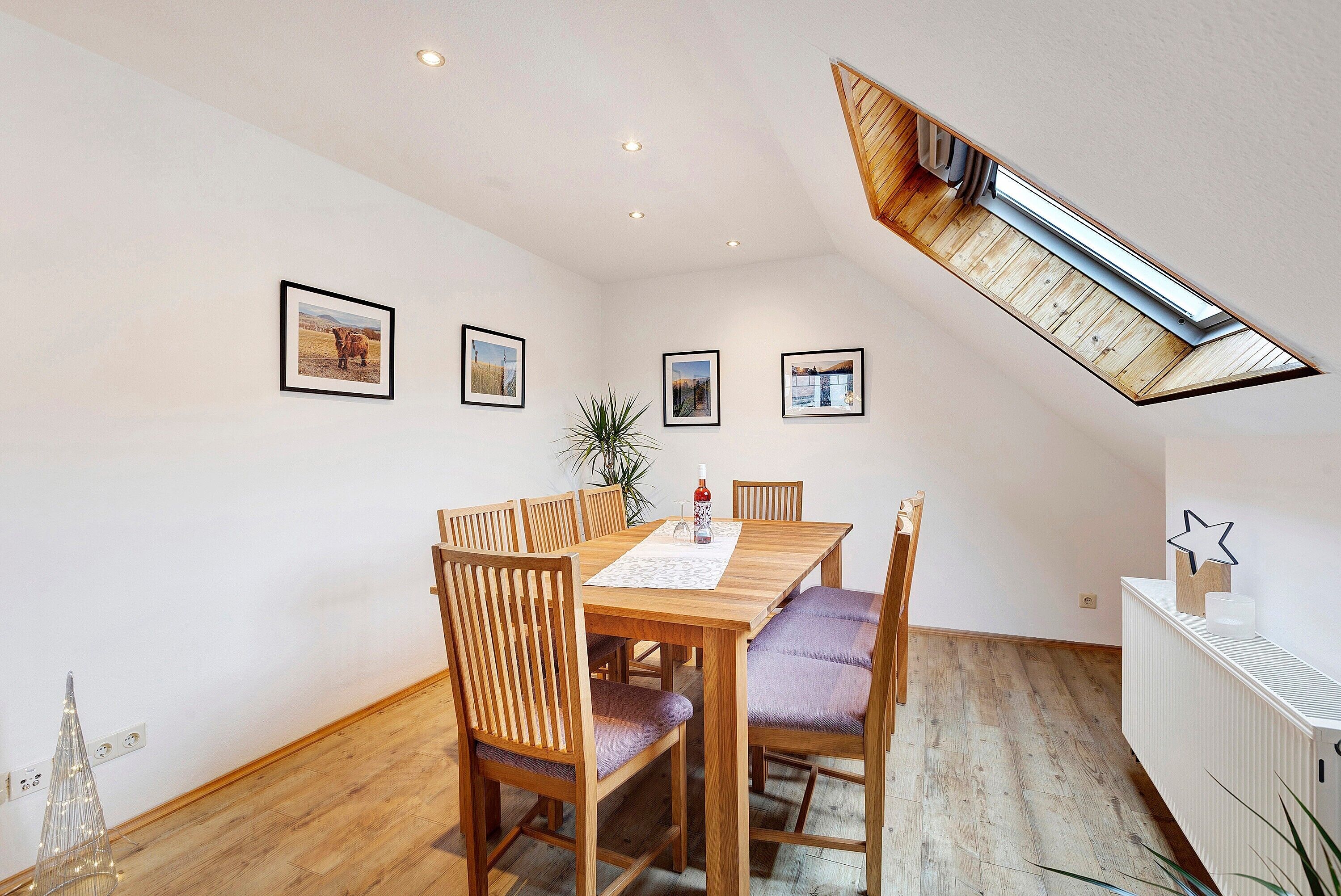 A dining area with a wooden table and chairs, a skylight, and framed pictures on the wall.