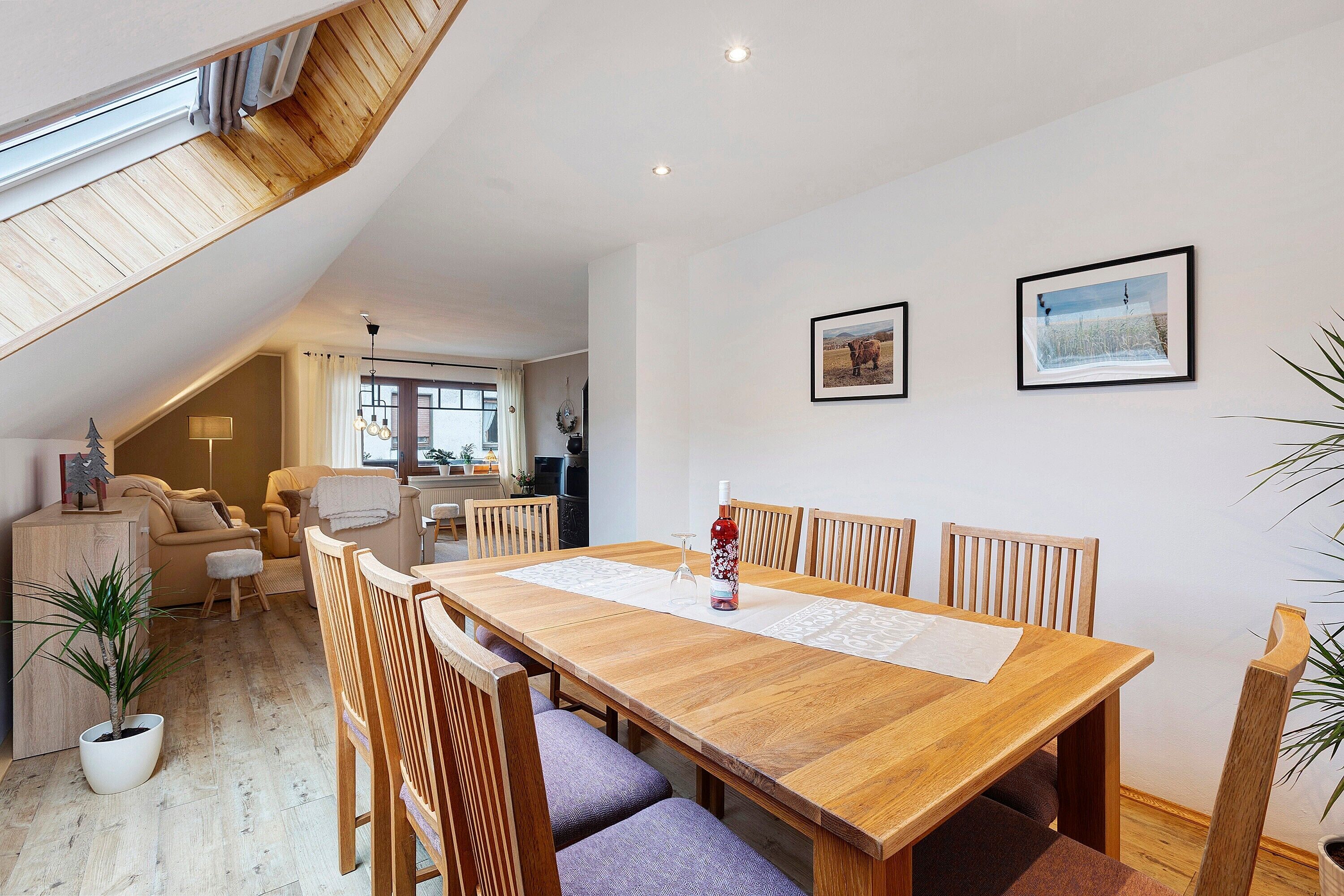 A dining area with a wooden table, chairs, and a bottle of wine. There are two framed pictures on the wall.
