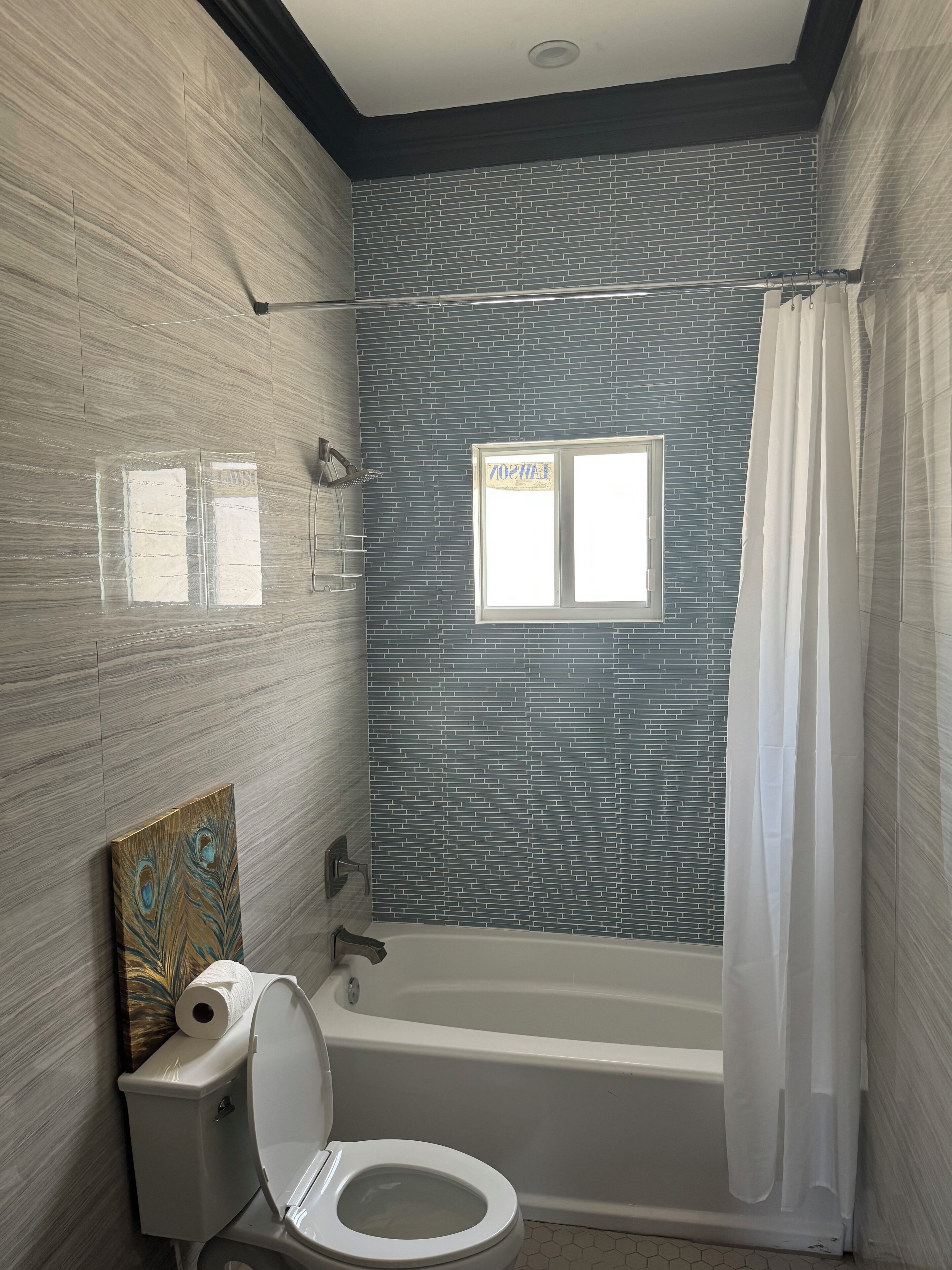 A bathroom with a peacock-themed artwork, a toilet, a bathtub, and a window with a view.