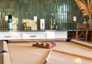 Une table de billard avec un queue et des billes, une chaise en bois et un vase contenant des plumes d’herbe des pampas.