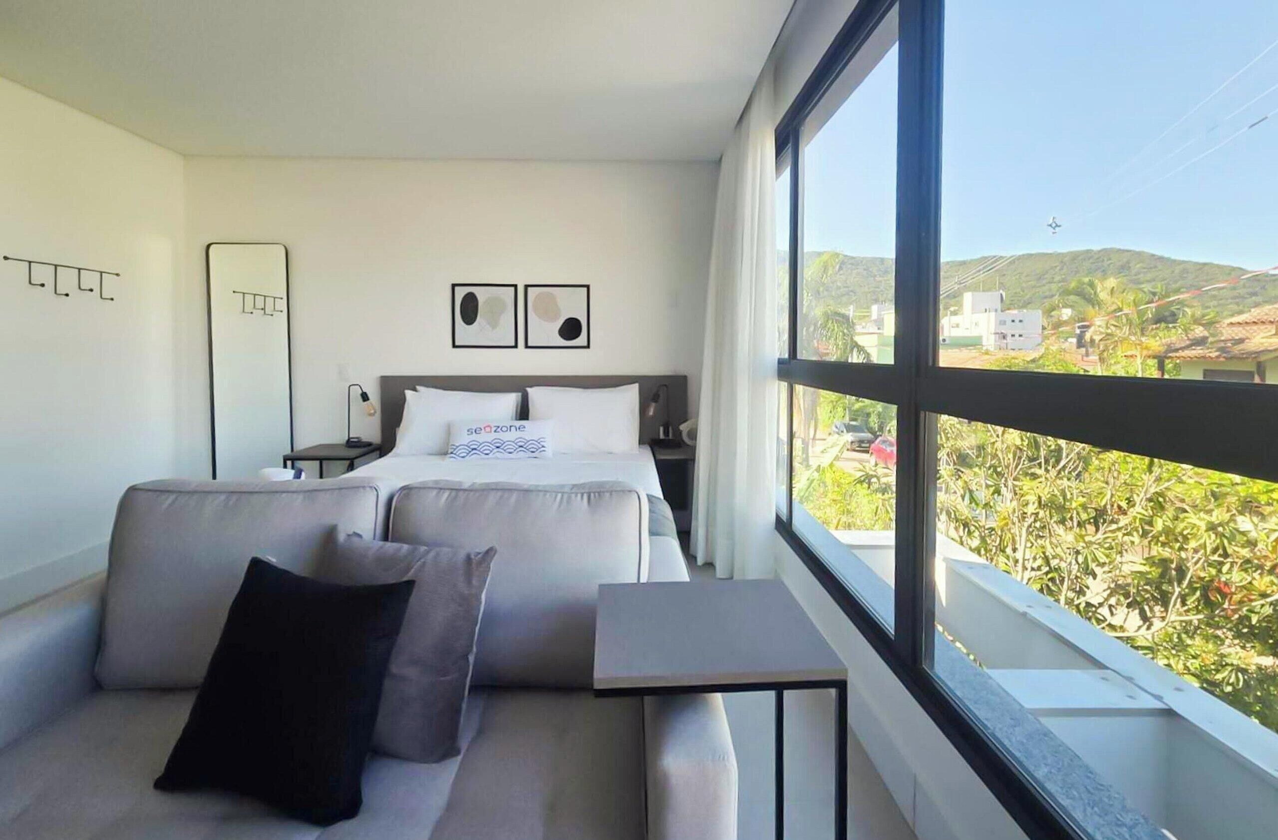 Quarto de hotel moderno com uma cama, um sofá, uma mesinha e uma janela ampla com vista para o exterior.