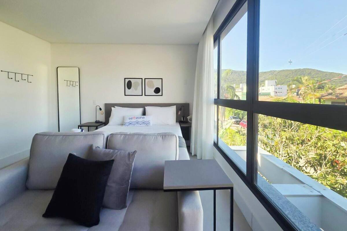 A modern hotel room with a bed, a sofa, a small table, and a large window offering a view of the outdoors.