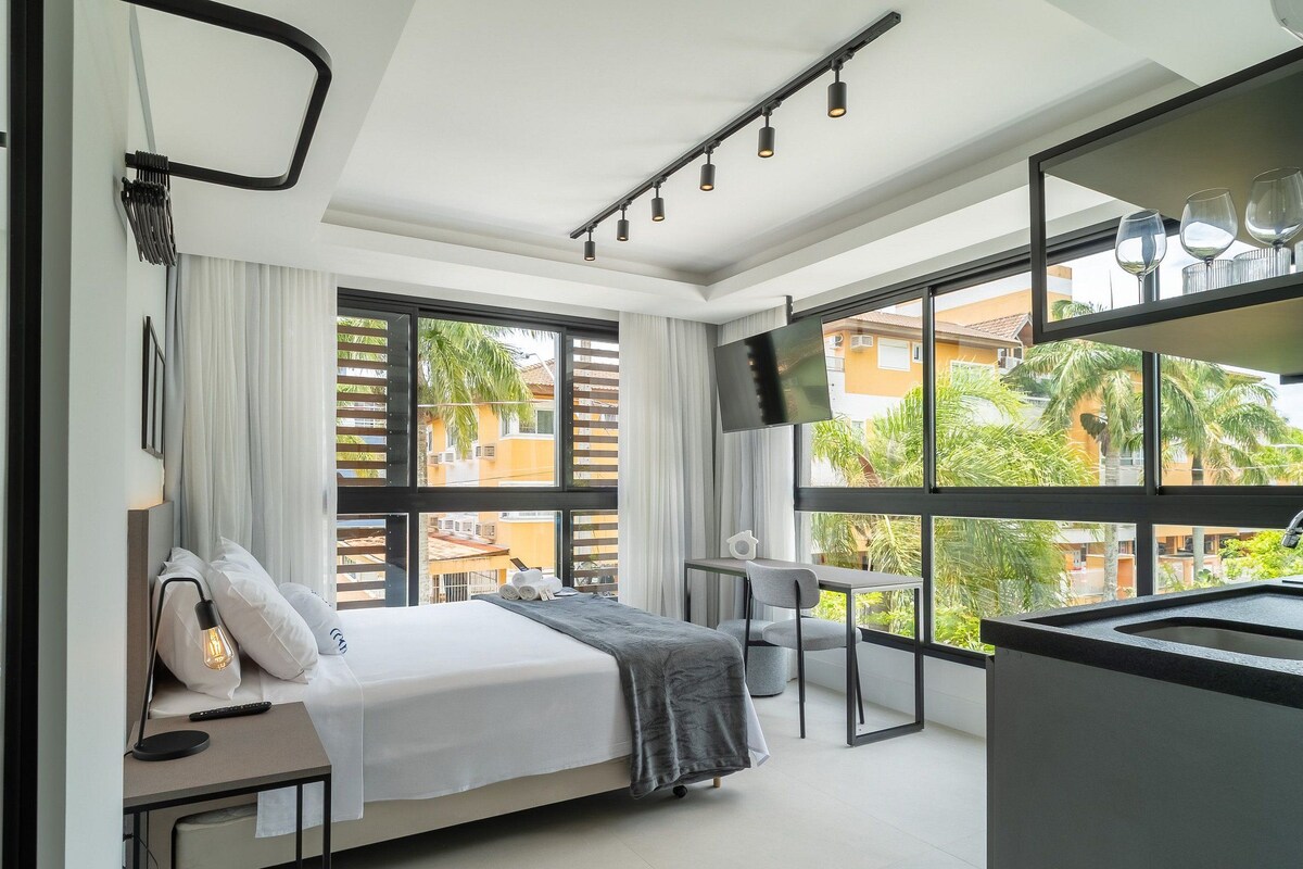 A modern hotel room with a bed, bedside table, lamp, and large windows offering a view of the outside.
