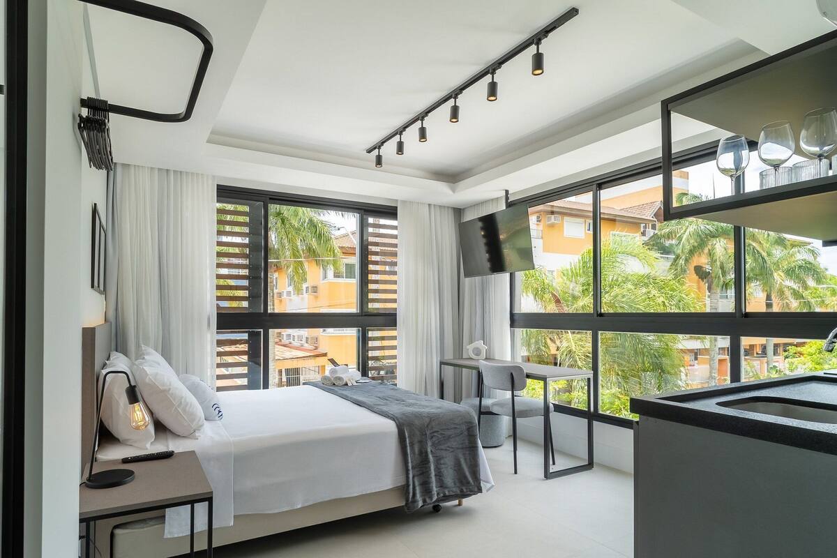 A modern hotel room with a bed, bedside table, lamp, and large windows offering a view of the outside.