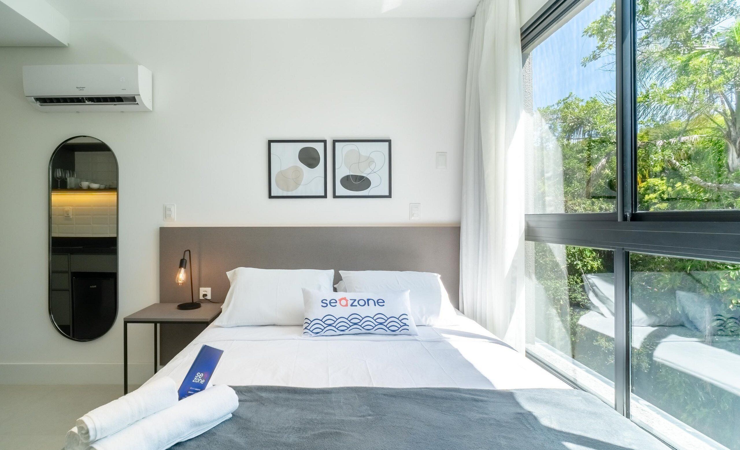 Quarto de hotel moderno com uma janela grande, cama com roupa de cama branca e almofada 'seazone', espelho redondo e ar-condicionado fixado na parede.