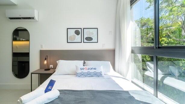 A modern hotel room with a large window, a bed with white bedding and a 'seazone' pillow, a round mirror, and a wall-mounted air conditioner. - Modern Studios 150m From Lagoa da Conceicao - Lsts I (Florianópolis)