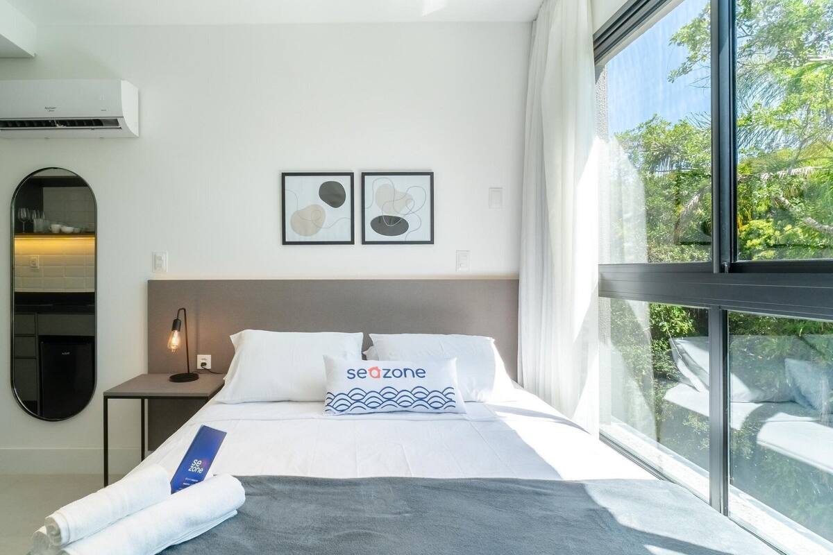 A modern hotel room with a large window, a bed with white bedding and a 'seazone' pillow, a round mirror, and a wall-mounted air conditioner.
