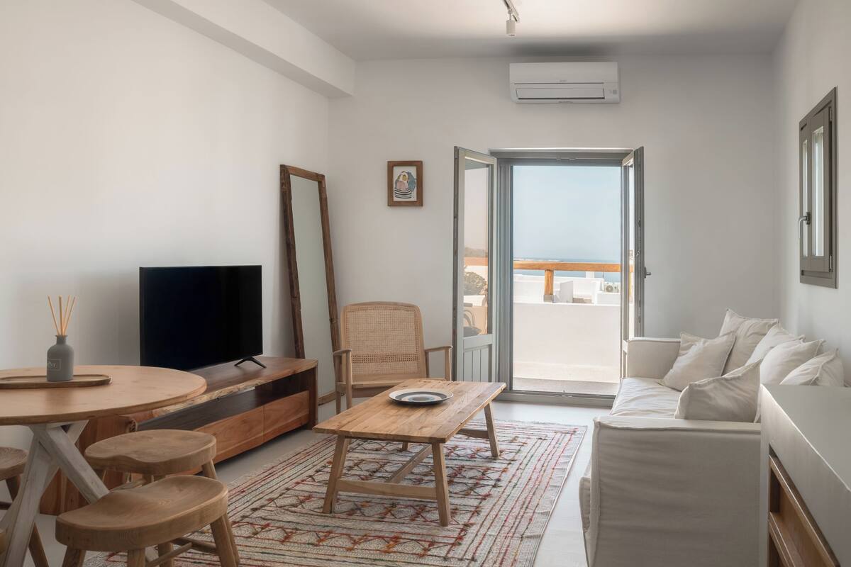 A modern living room with a flat-screen TV, wooden furniture, a dining area, and a balcony with a view.