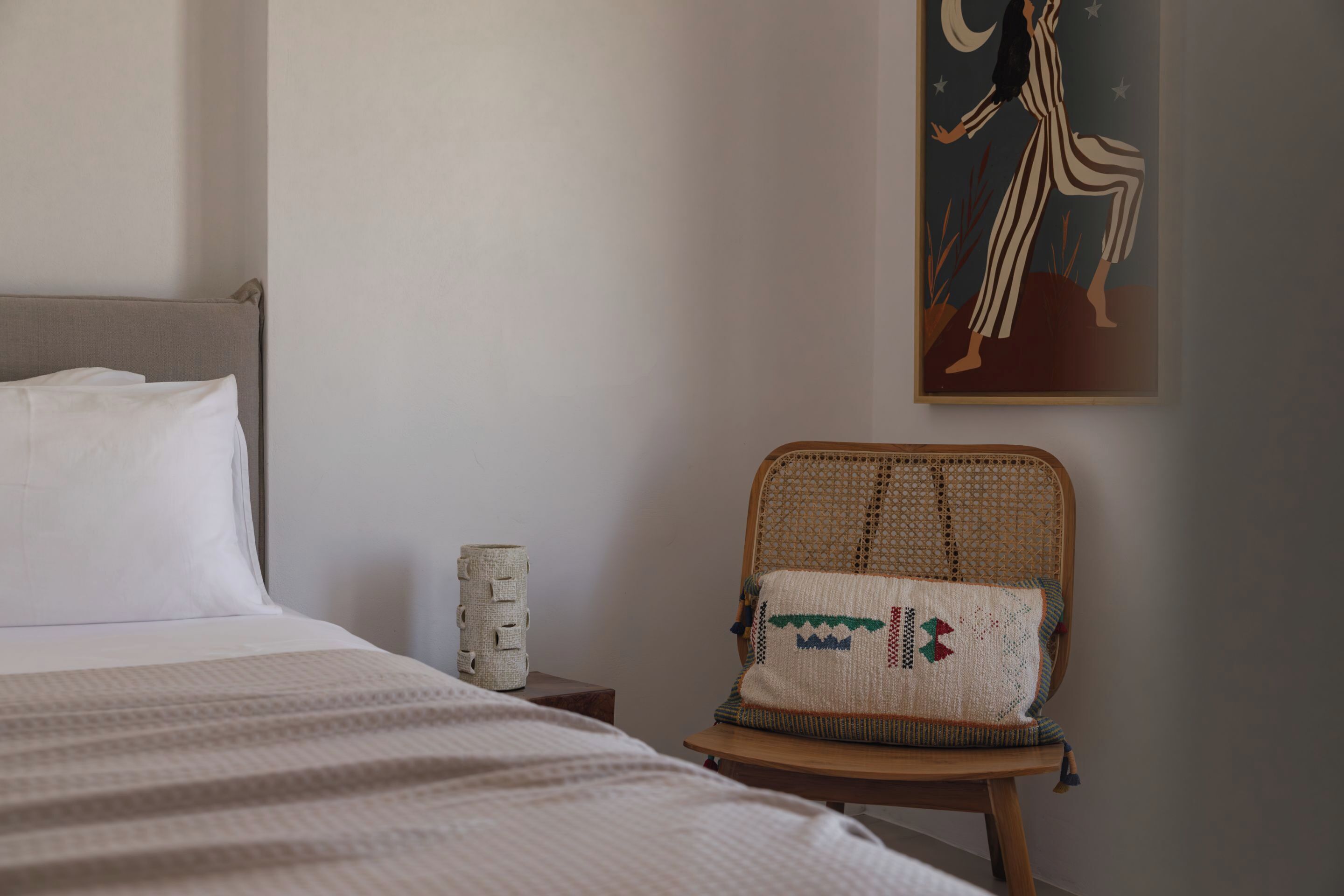 A neatly made bed with white linens, a woven chair with a patterned cushion, and a wall-mounted painting of a woman in a striped outfit.