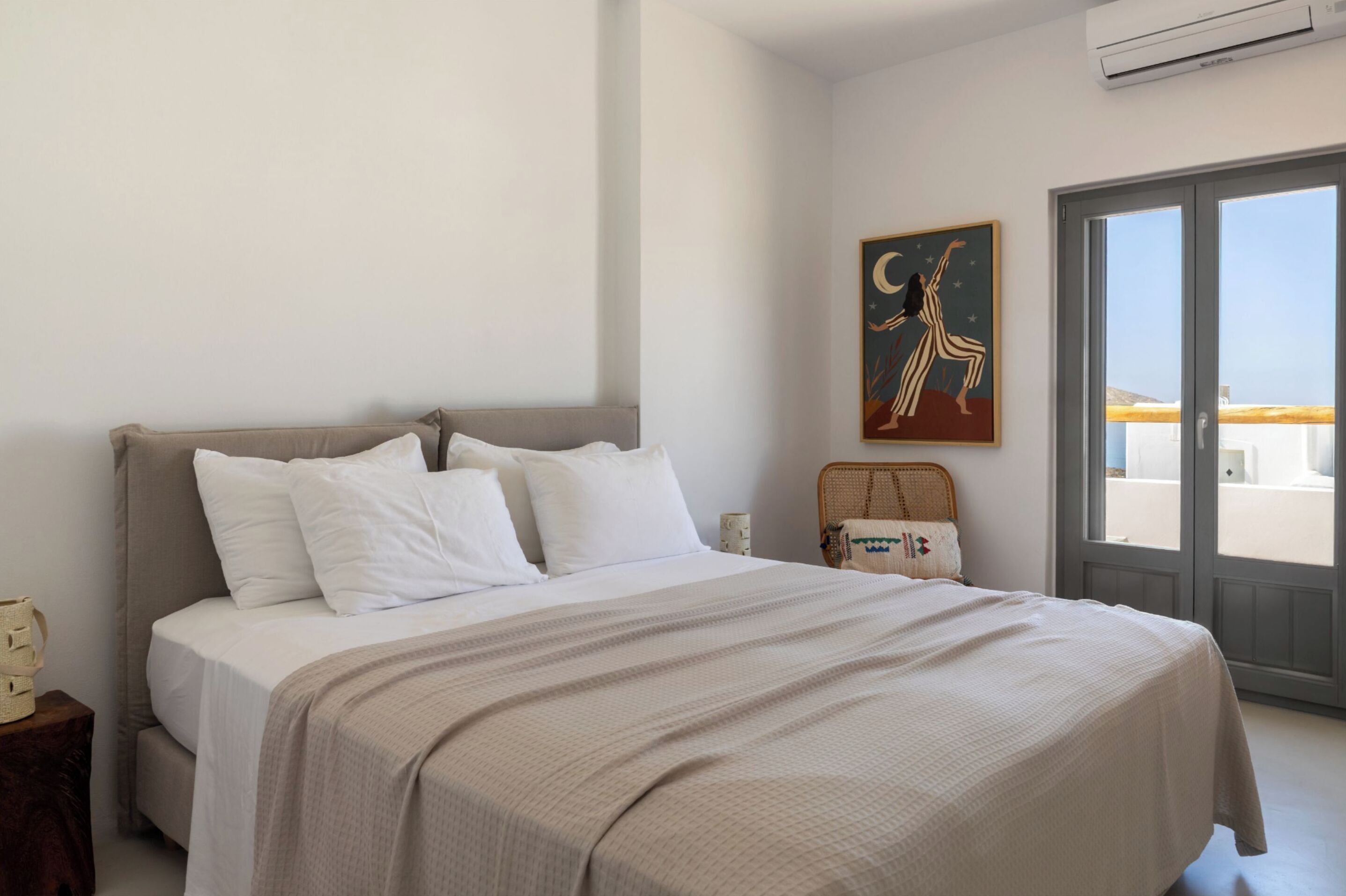 A bedroom with a bed, a nightstand, a wicker chair, a wall-mounted artwork, and a sliding glass door leading to a balcony.