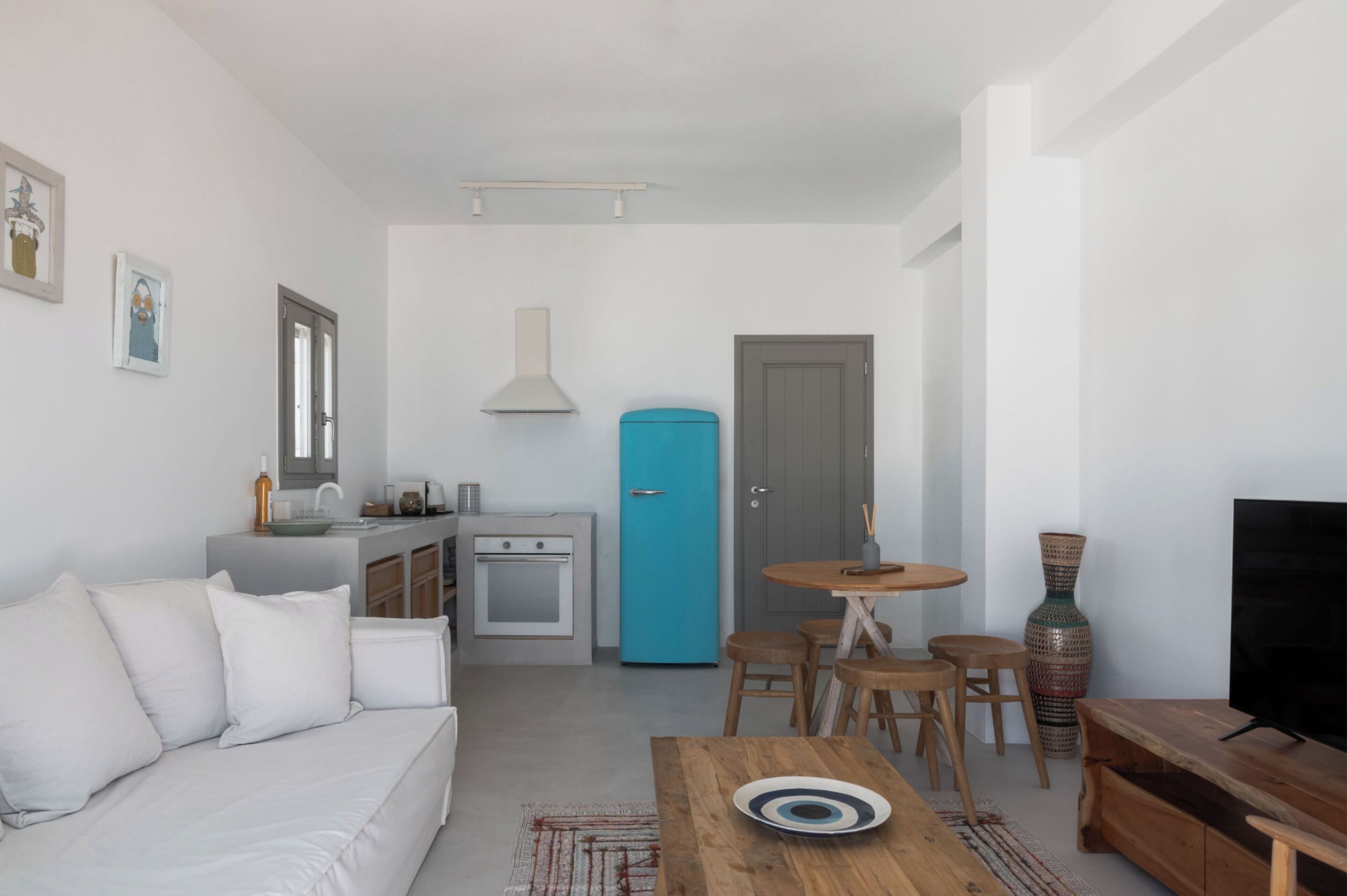 A modern living room with a white sofa, a wooden coffee table, and a TV. There is a kitchen area with a blue refrigerator and a stove.