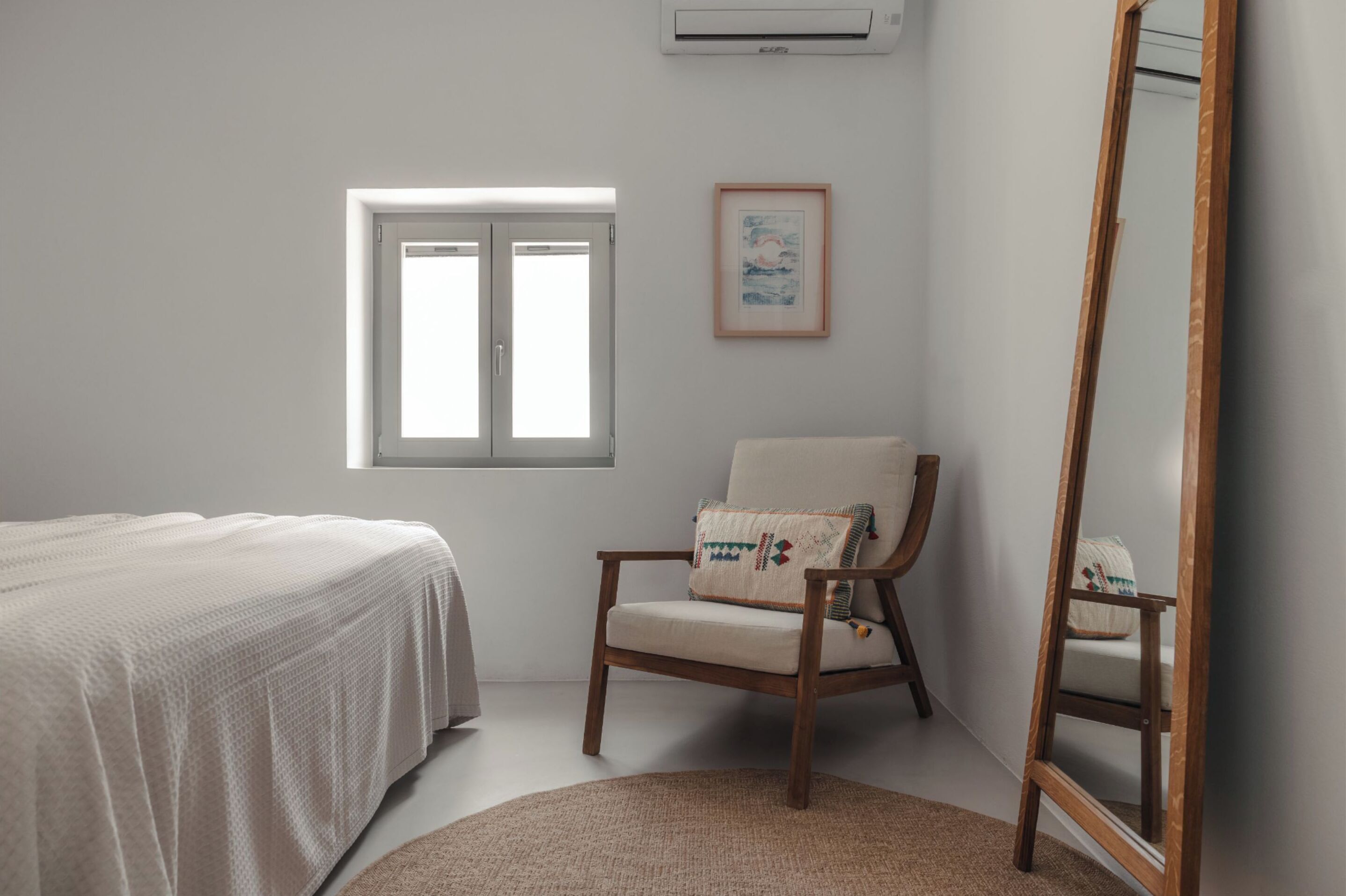 A bedroom with a bed, a chair, a window, a framed picture, and a mirror.