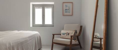 A bedroom with a bed, a chair, a window, a framed picture, and a mirror.