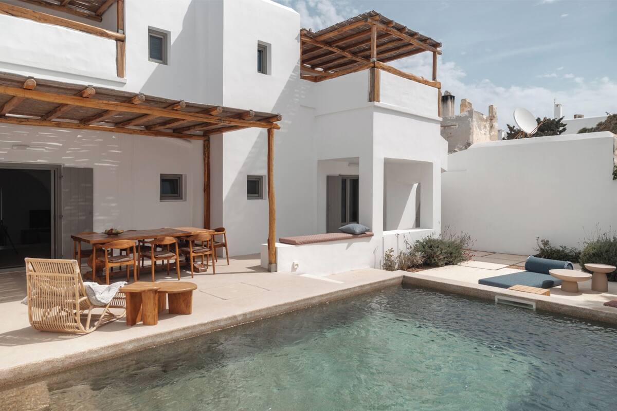 A modern outdoor pool area with a wooden pergola, a dining set, and a seating area with cushions.
