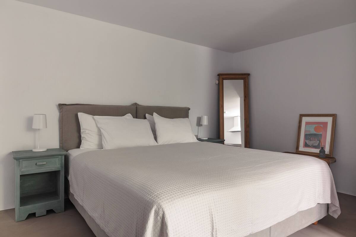 Un lit bien fait, avec une couette blanche et des oreillers, une petite table de chevet avec une lampe, un miroir et un tableau encadré au mur.
