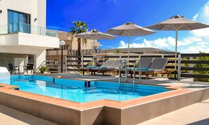 A modern outdoor swimming pool with lounge chairs and umbrellas, surrounded by a multi-level building with glass railings. - Pnoe Beachside Suite (Georgioupoli)