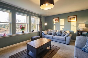 A living room with a grey sofa, a wooden coffee table, and a rug. There are two windows with curtains, a lamp, and framed pictures on the wall. - Host Stay The Annex (Jedburgh)