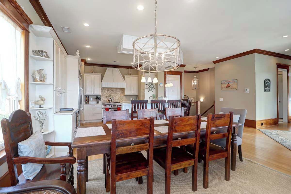 A dining area with a wooden table and chairs, a chandelier, and a kitchen in the background.