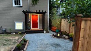 A residential driveway leading to a house with a red door, a wooden fence, and a garden area with potted plants.