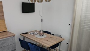Een moderne eethoek met een houten tafel, twee blauwe stoelen, een zwart aan de muur bevestigd televisietoestel en een hanglamp.