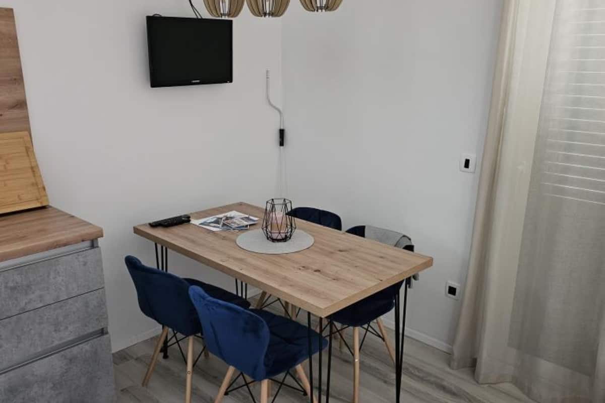 Un espace repas moderne avec une table en bois, deux chaises bleues, un téléviseur noir fixé au mur et un luminaire suspendu.