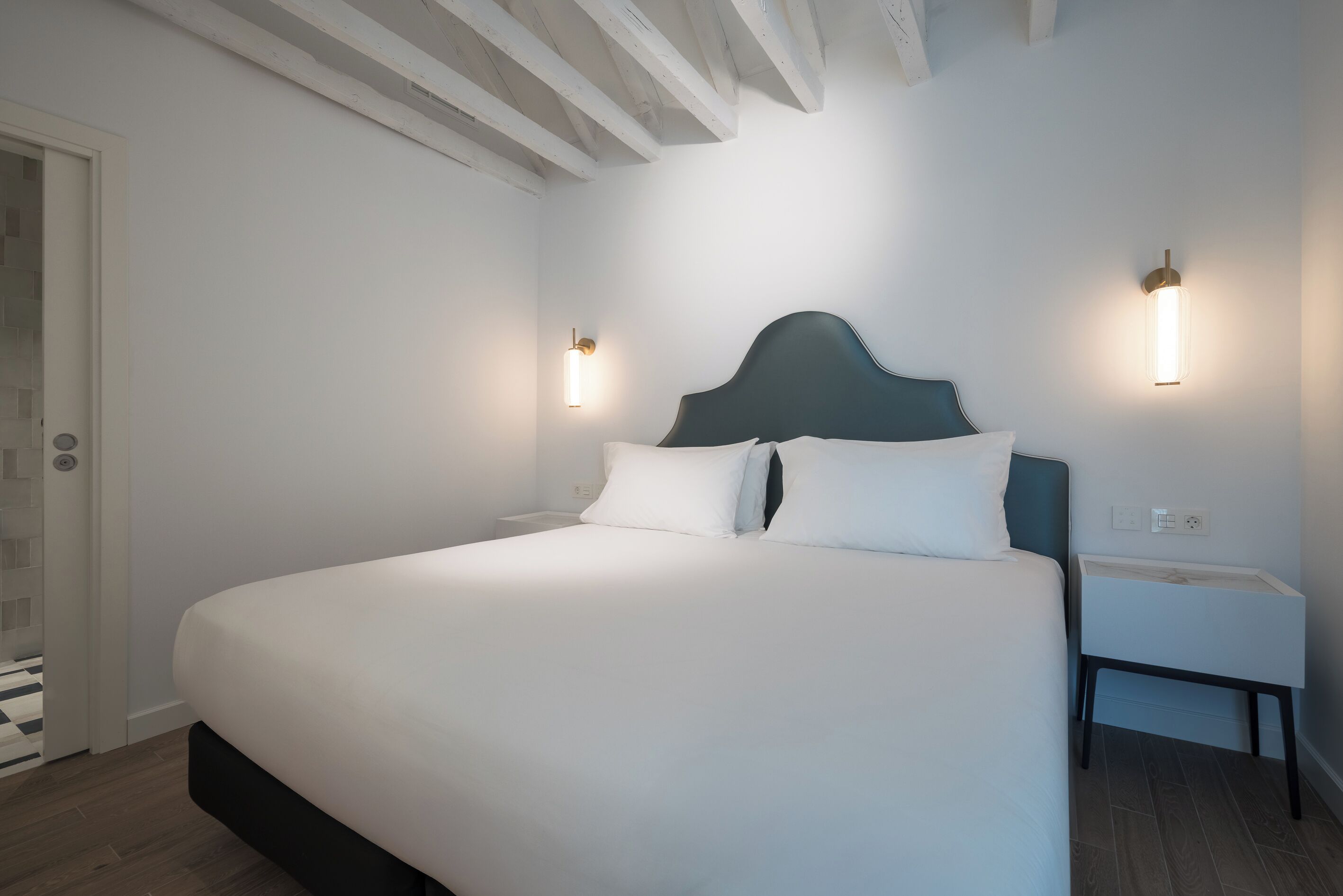 Un lit bien fait, avec une tête de lit sombre, dans une pièce aux murs blancs et aux poutres apparentes au plafond. On y trouve deux tables de chevet, chacune équipée d’une lampe fixée au mur.