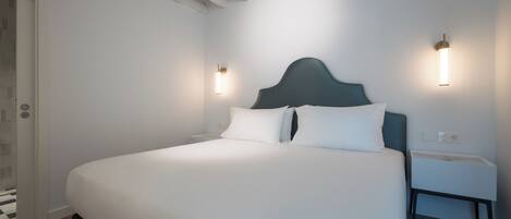 A neatly made bed with a dark headboard in a room with white walls and wooden beams on the ceiling. There are two bedside tables with wall-mounted lamps.