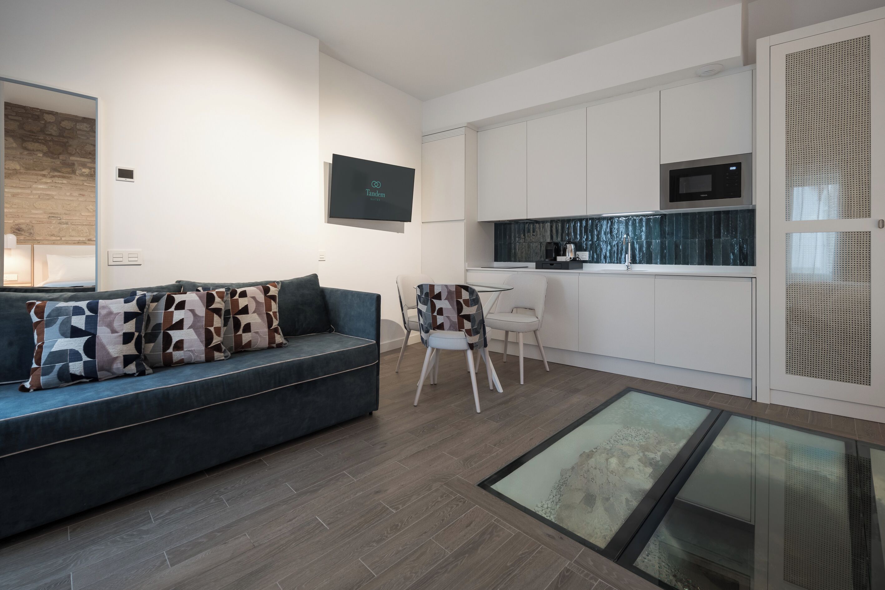 A modern living room with a sofa, a dining table, and chairs. There is a kitchen area with white cabinets and a microwave.