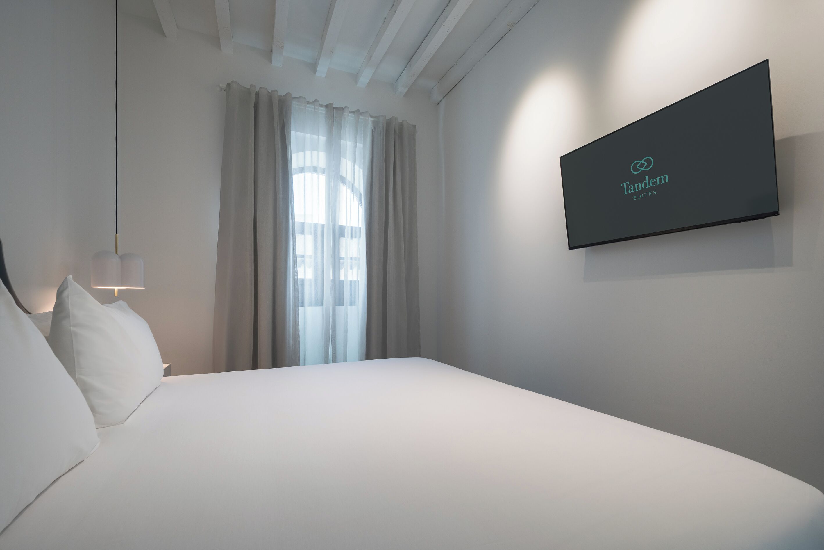 A modern hotel room with a large bed, a mounted TV displaying 'Tandem Suites', and a window with sheer curtains.