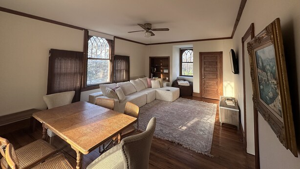Amplia sala de estar con mesa de comedor de madera, asientos acolchados, ventanas grandes y un ventilador de techo.