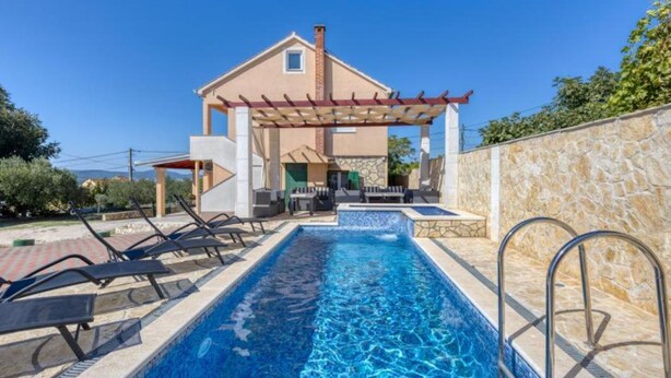 Une maison à deux étages avec une piscine, un espace salon extérieur et un mur en pierre.