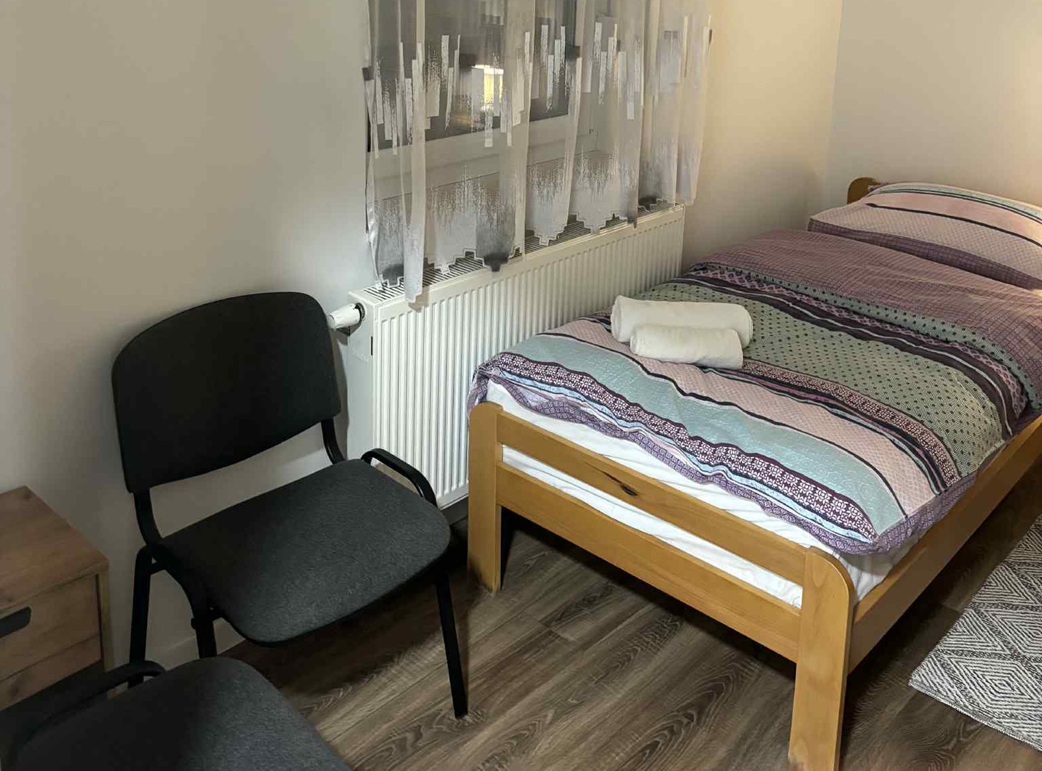 Un dormitorio con cama, una silla, un radiador y una ventana con cortinas translúcidas.