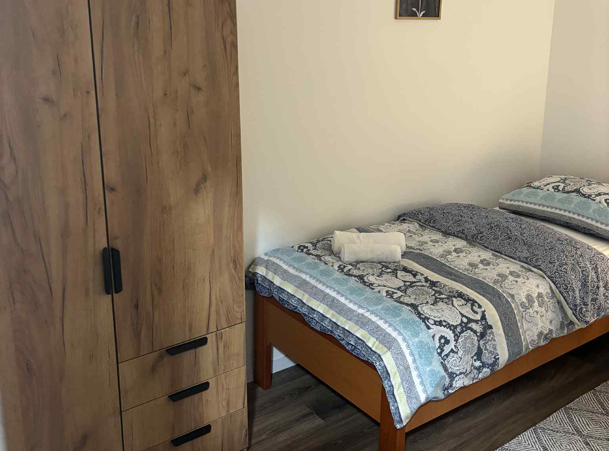 A bedroom with a wooden wardrobe, a bed with patterned bedding, and a small wall-mounted shelf.