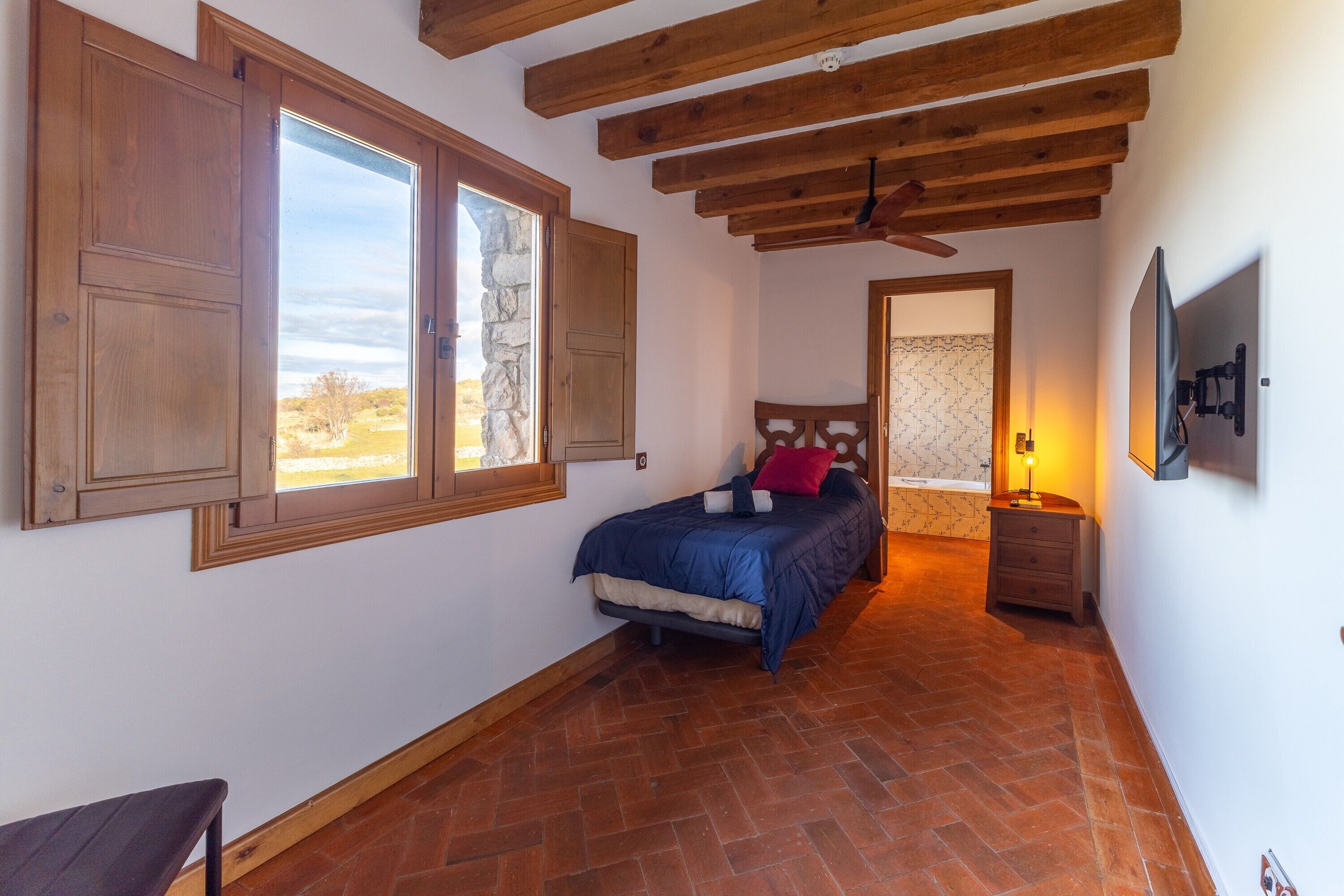 Une chambre à coucher avec un lit, des poutres apparentes en bois au plafond, une fenêtre, une table de chevet et un téléviseur fixé au mur.