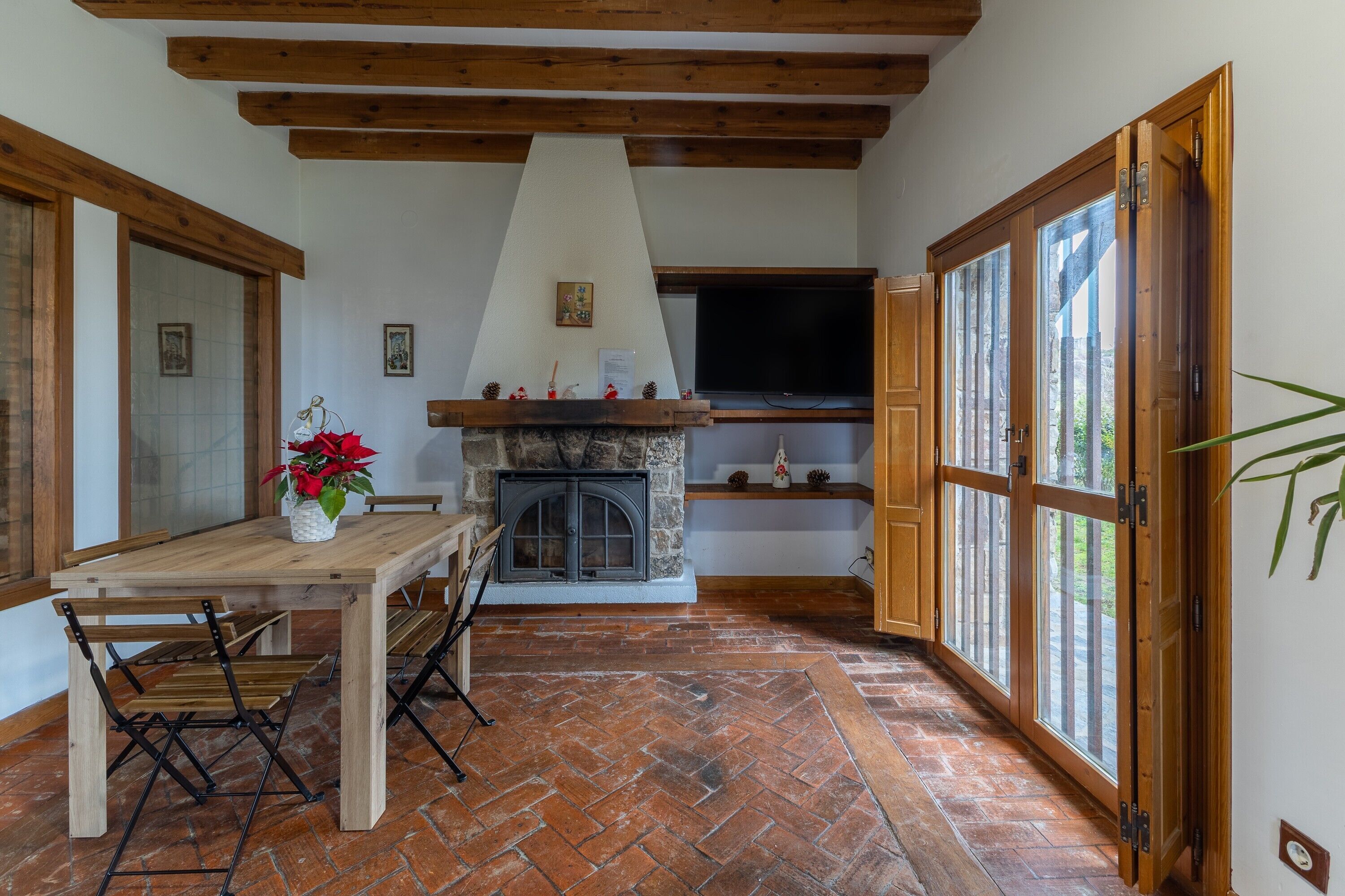 Una habitación con chimenea de piedra, vigas de madera en el techo, una mesa de comedor con sillas y ventanas grandes.