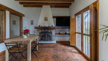 Una habitación con chimenea de piedra, vigas de madera en el techo, una mesa de comedor con sillas y ventanas grandes.