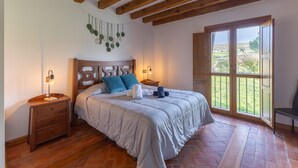 Un dormitorio con cabecera de madera, una cama con ropa de cama blanca, una mesita de noche con lámpara y una ventana con vistas a zonas verdes.
