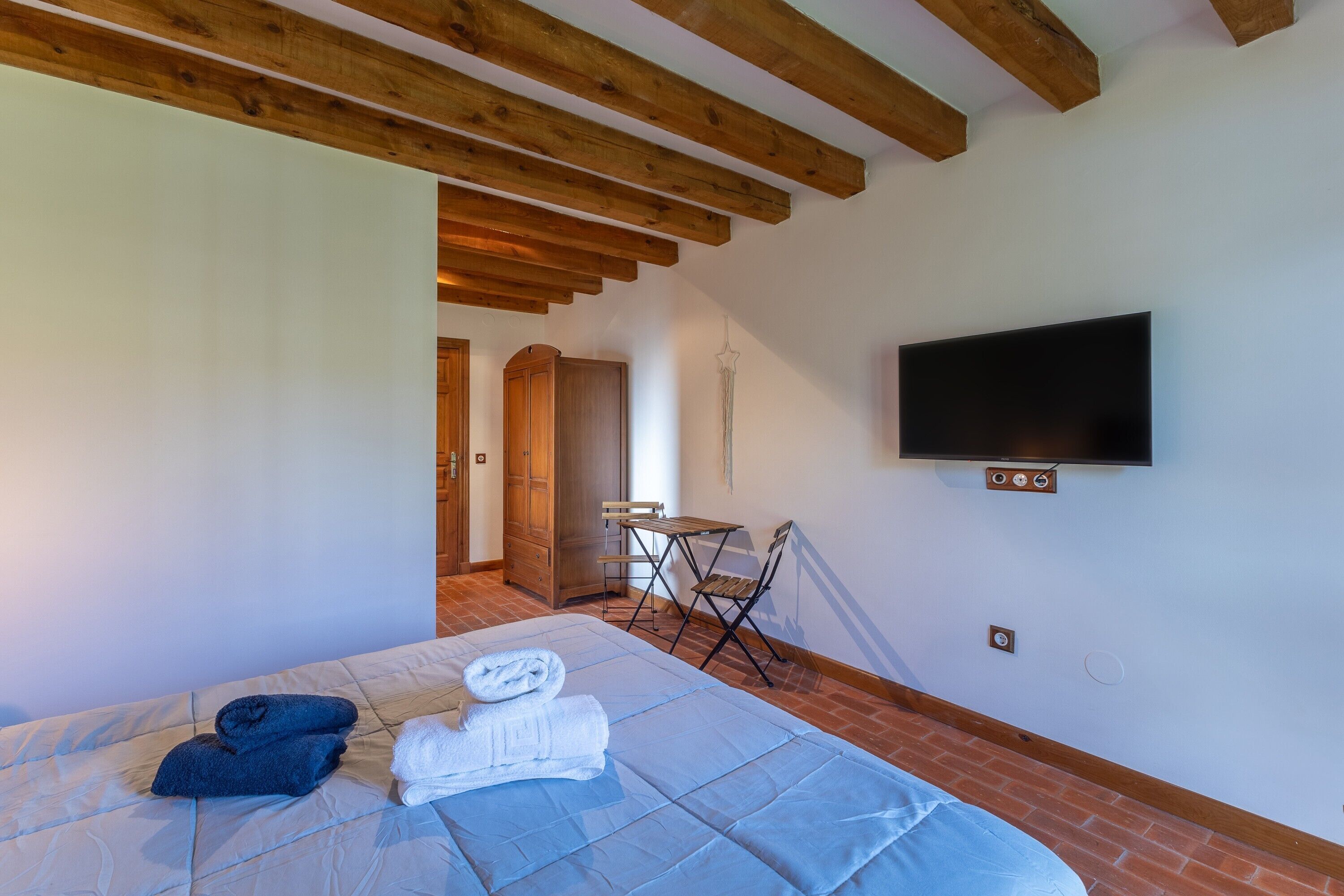 Un dormitorio con una cama, televisor montado en la pared, techo de madera y suelo de ladrillo.
