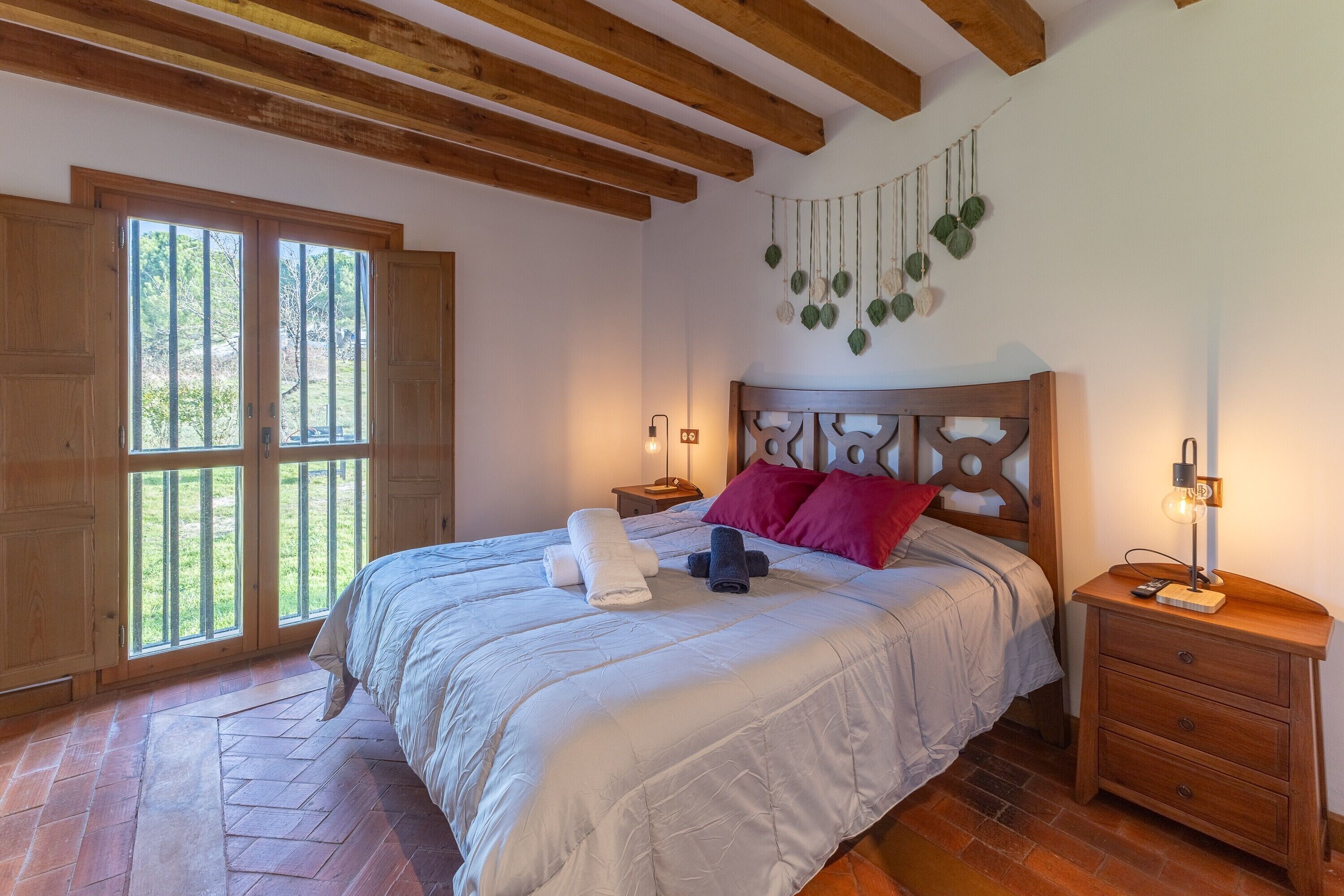 Une chambre à coucher avec un lit, une tête de lit en bois, des tables de chevet, une plante suspendue et une fenêtre donnant sur un beau paysage.