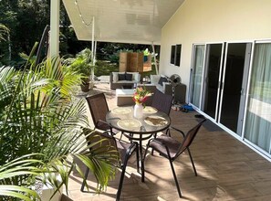 A covered patio with a round table, chairs, and a fan. There are potted plants and a sofa with cushions.