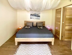 A bedroom with a wooden bed, two pillows, a nightstand with a lamp, a wall-mounted painting, and a door.
