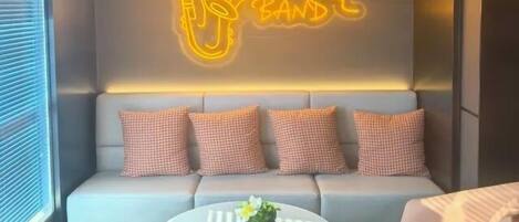 A modern living area with a neon sign reading "JAZZ BAND" on the wall, a sofa with patterned cushions, and a round glass table with a flower centerpiece.