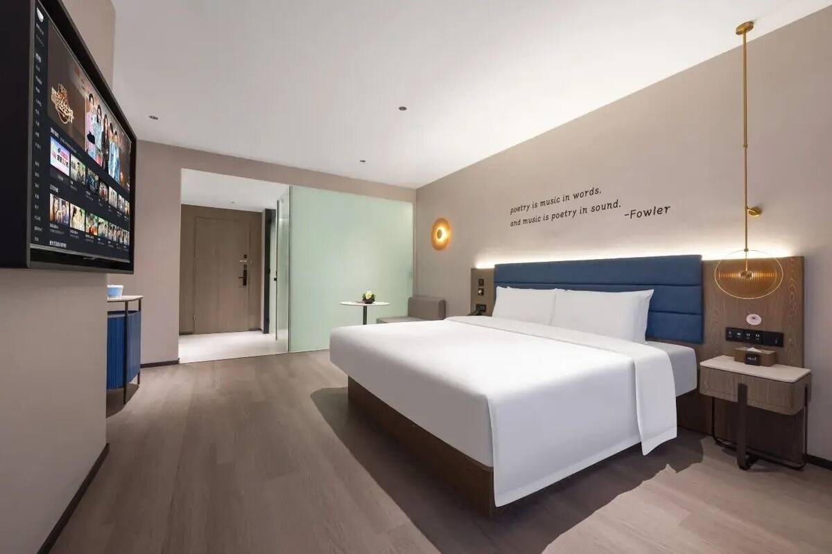 Une chambre d’hôtel moderne avec un grand lit, un téléviseur à écran plat fixé au mur, une table de chevet avec une lampe et une citation affichée sur le mur.